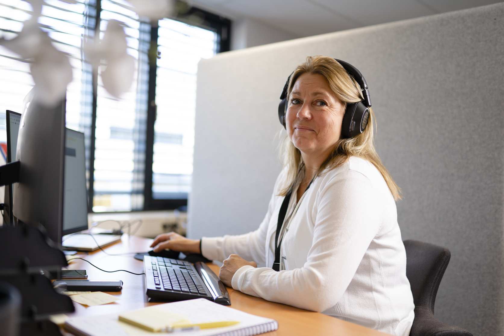 Kvinne med hodetelefoner sitter ved en datamaskin på et kontor, ser opp fra skjermen og smiler svakt. På pulten ligger notatbok og tastatur